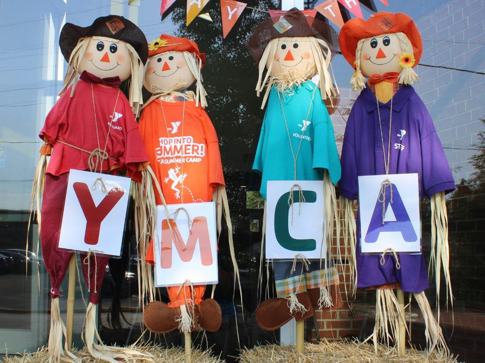 Four scarecrows wearing YMCA t-shirts hold signs for the letters Y, M, C, and A.